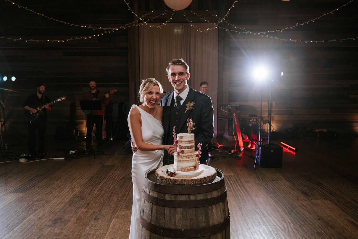 Couple cuts their wedding cake displayed on rustic wooden barrel under string lights.
