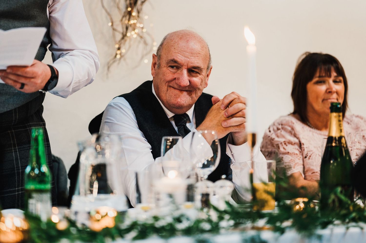 Wedding guests enjoying drinks and conversation at a decorated reception table with wine bottles.
