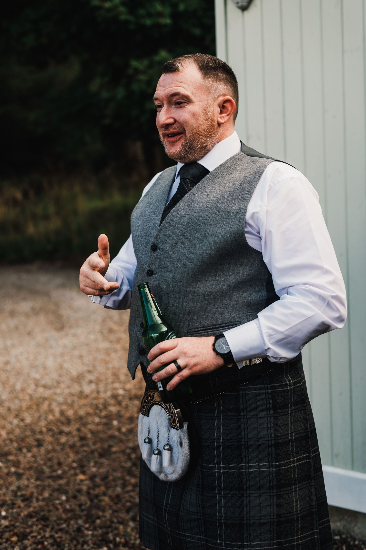 Wedding party members in kilts and vests enjoying drinks outdoors.