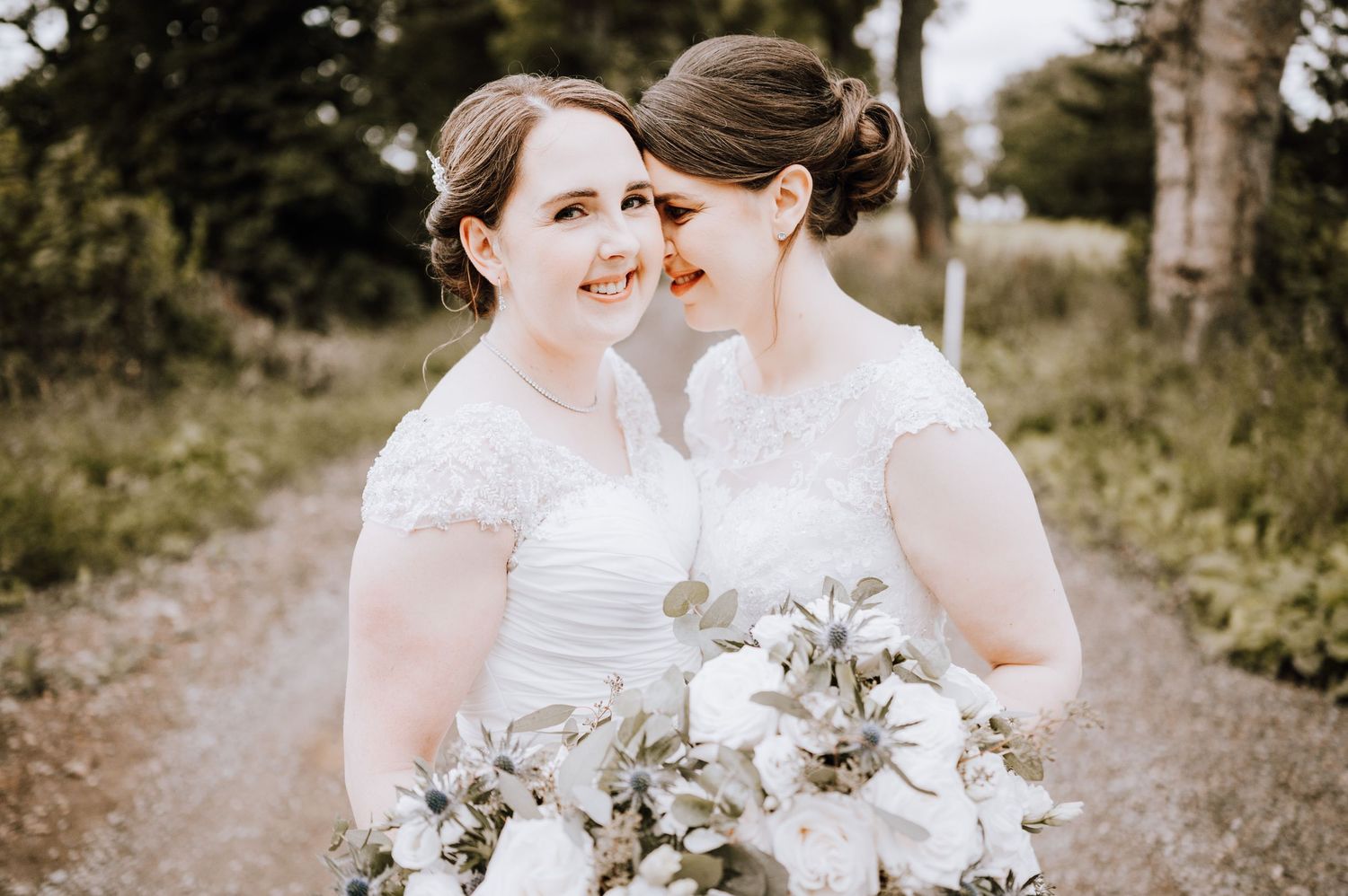 Two people in white wedding dresses share a joyful moment together outdoors surrounded by greenery. Meldrum House Wedding Photography.