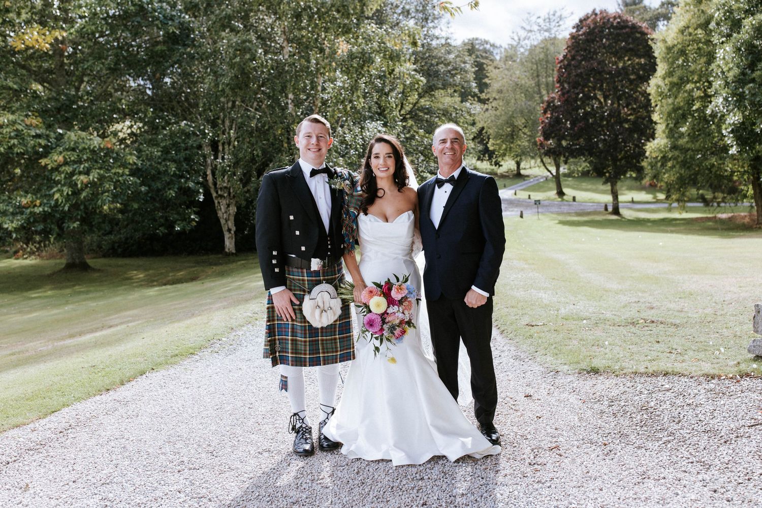 Three people in wedding attire including Scottish kilt stand together on garden path.