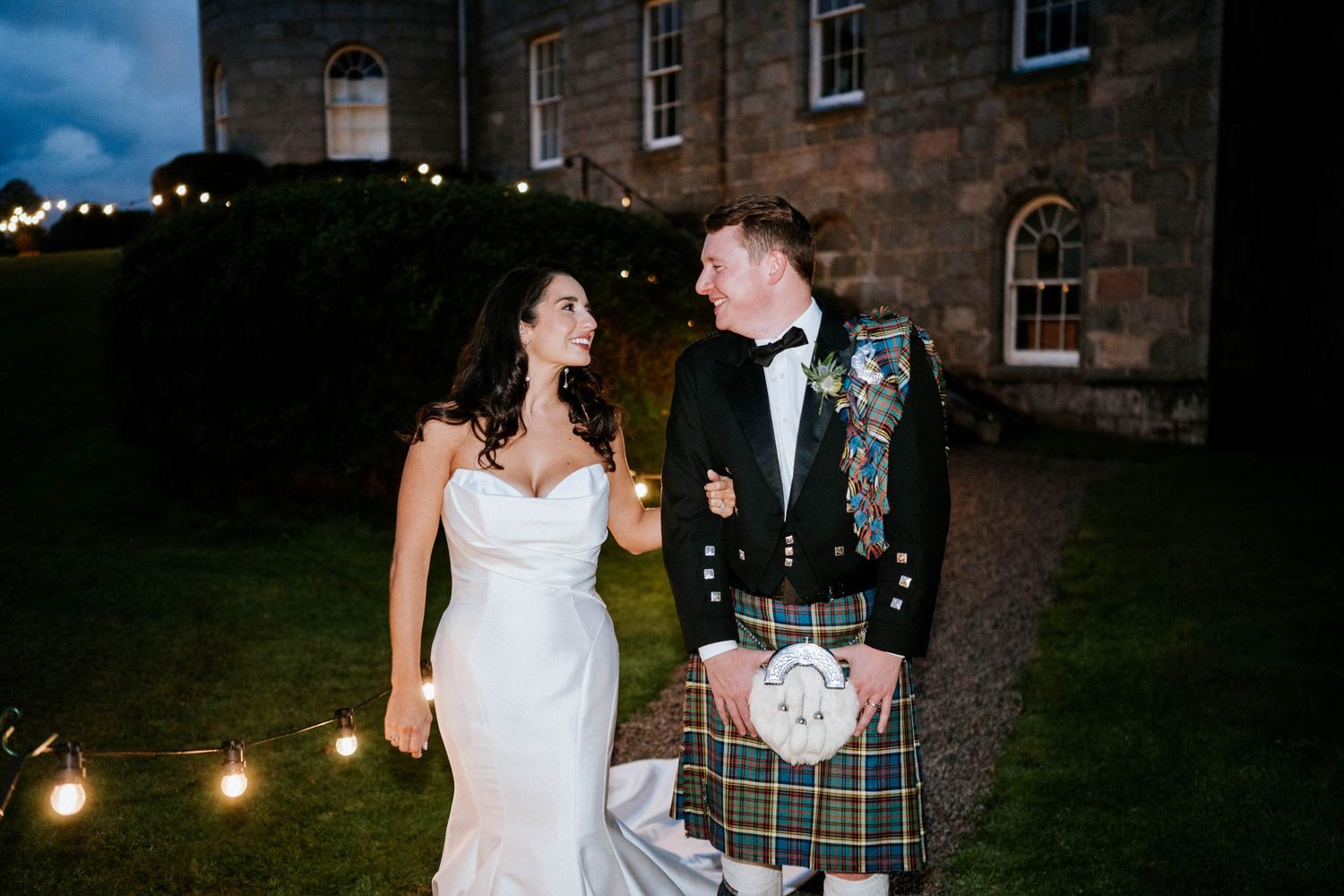 A bride in white gown and groom in traditional Scottish kilt pose together outside a stone building.
