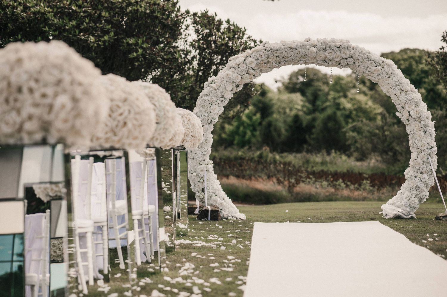 White floral wedding arch with scattered petals along aisle runner for outdoor ceremony.