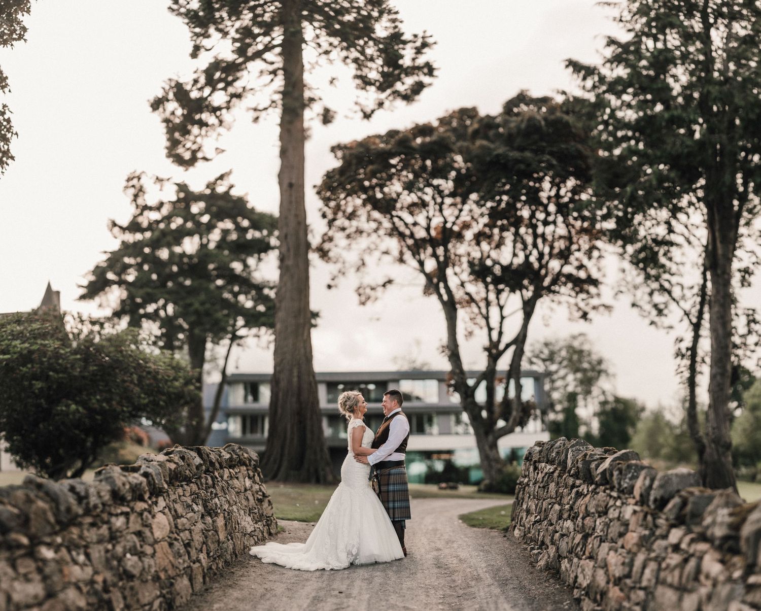 Wedding couple stands on stone-lined path with trees and buildings in background.