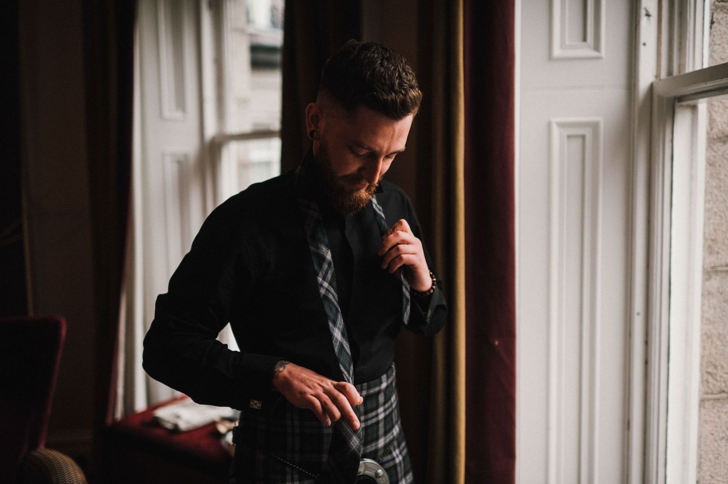 Figure in dark clothing and plaid kilt standing by a bright window in an elegant room.