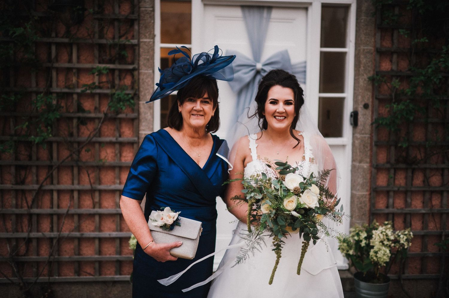Two wedding attendees posing in front of a brick building with decorative white bow.