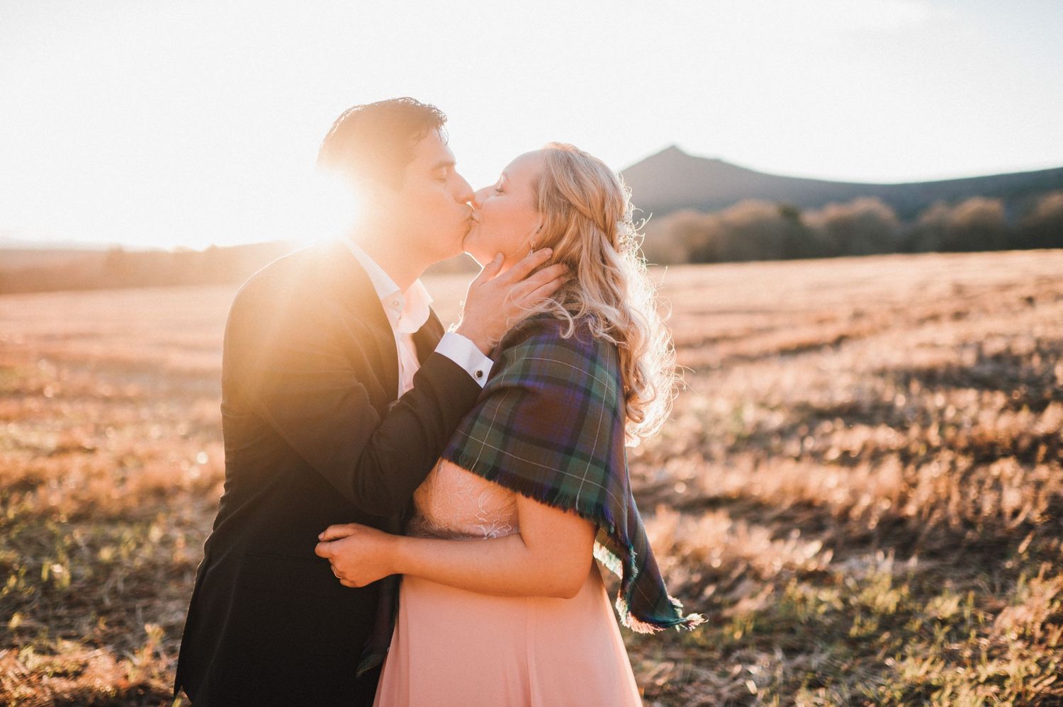 A couple shares a romantic kiss in a sun-drenched field with mountains in the background.
