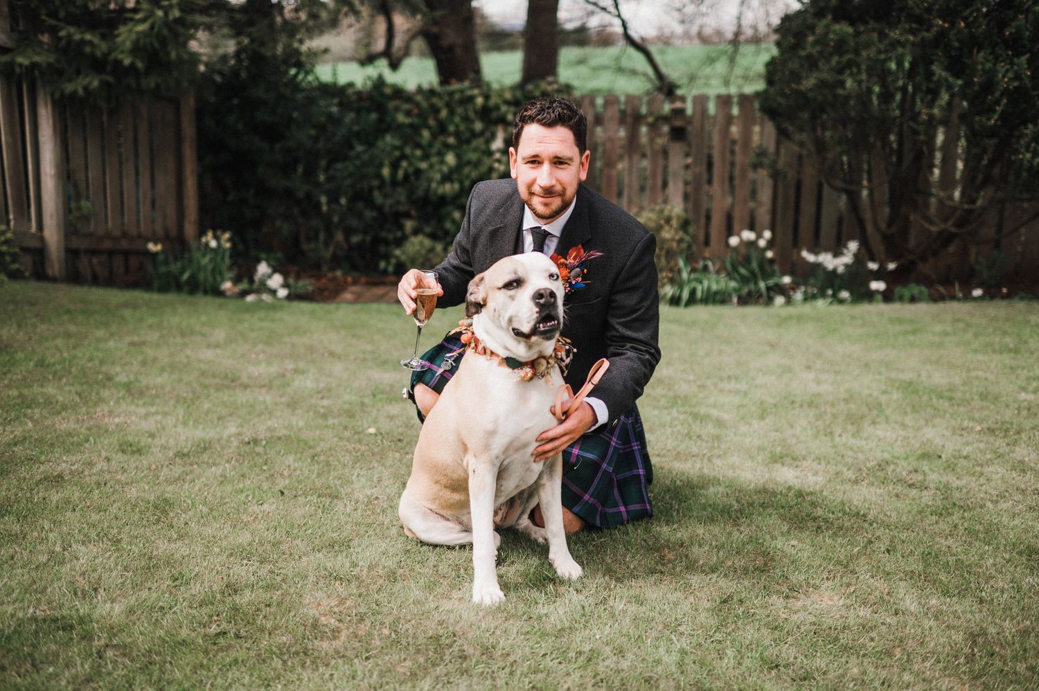 A happy light-colored dog wearing a festive collar sits with someone in formal attire on a green lawn.