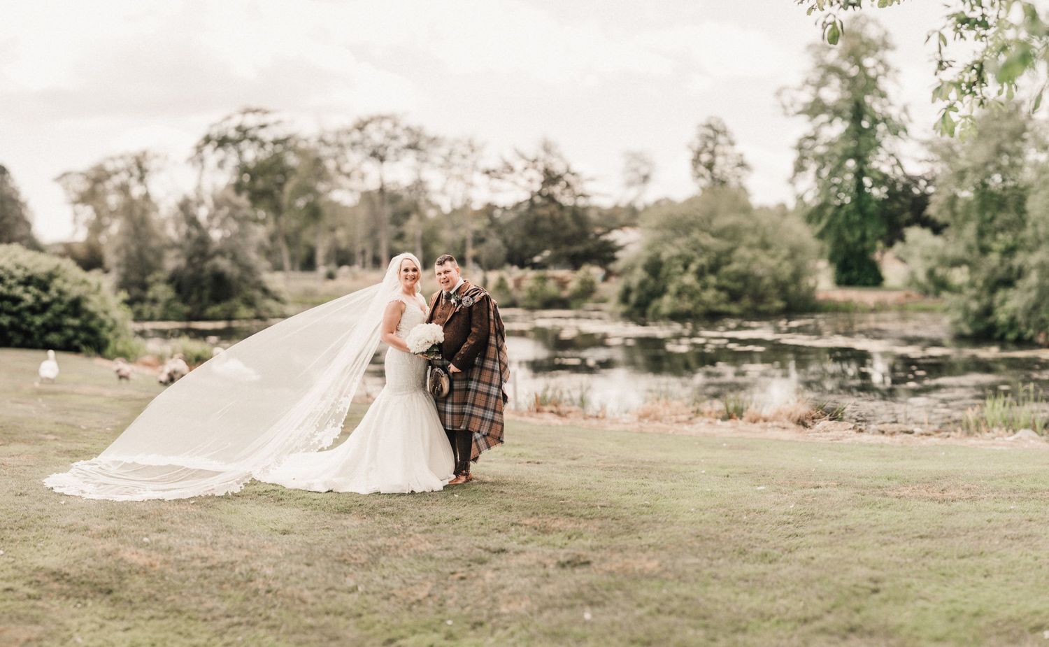 A wedding couple stands on manicured grounds with a scenic pond and trees in the background.