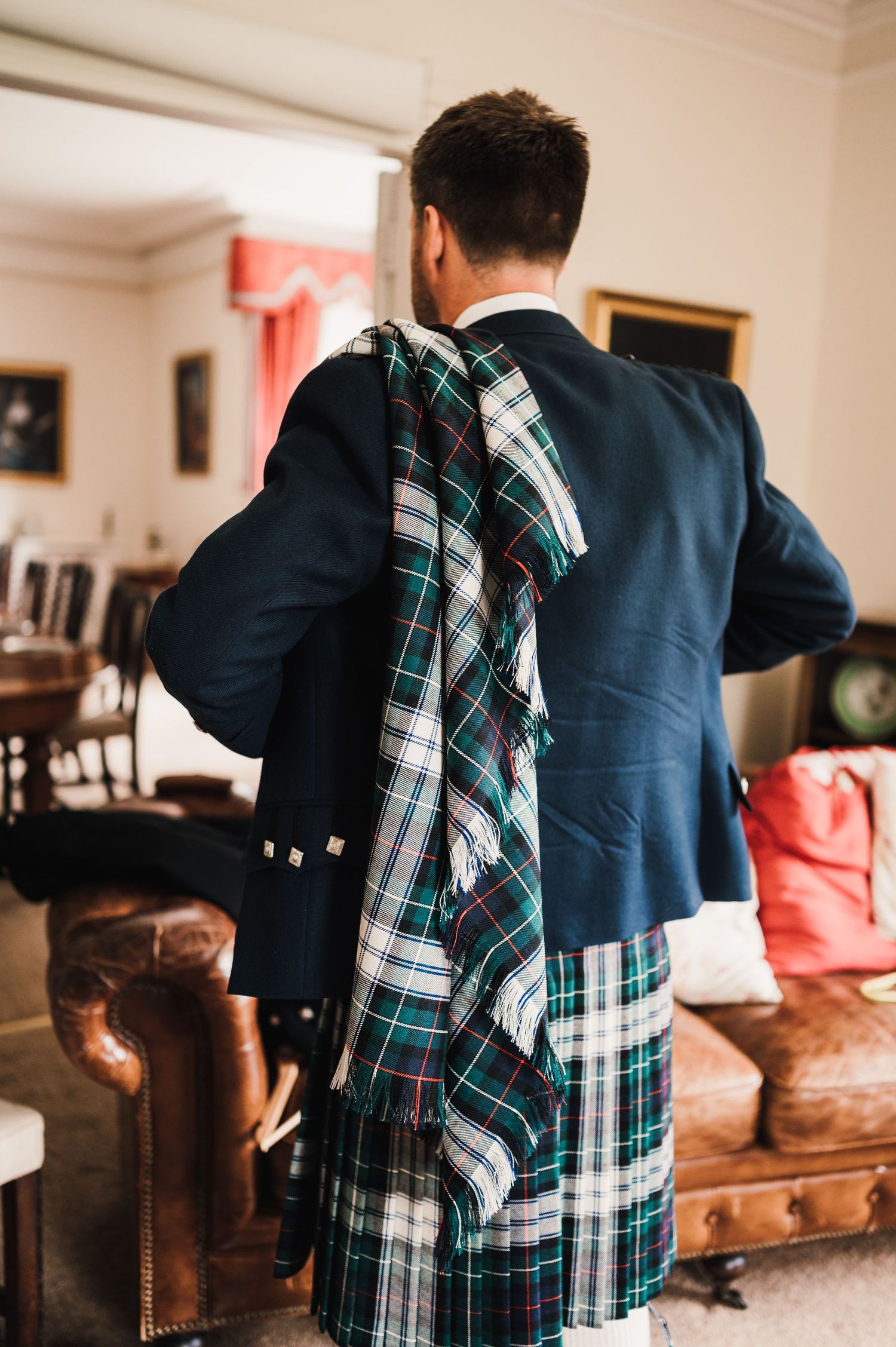 Man wearing traditional Scottish formal attire adjusts his tartan plaid against a warm interior setting.