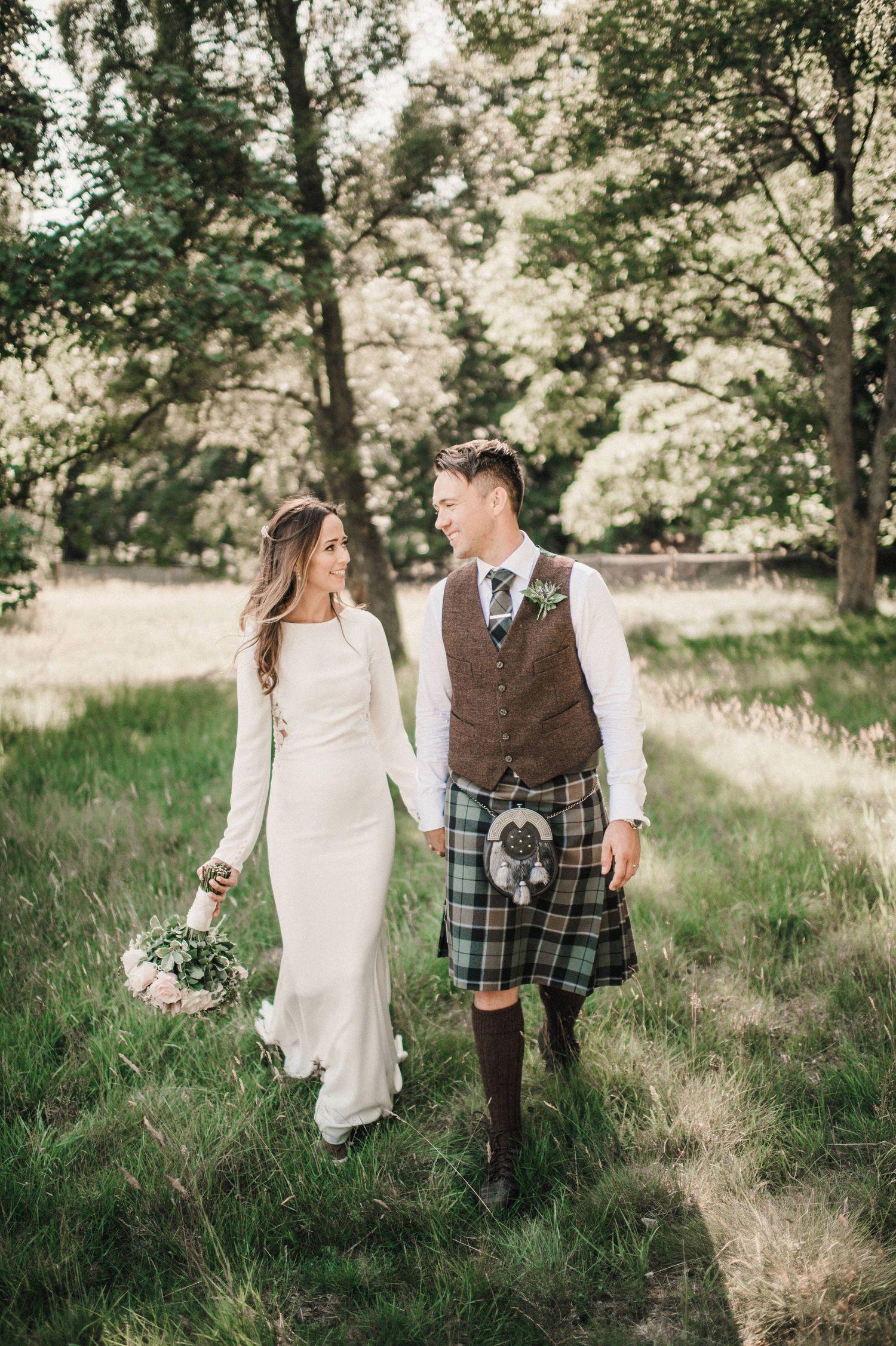 Bride in elegant white dress walks with kilted groom through a peaceful woodland setting.