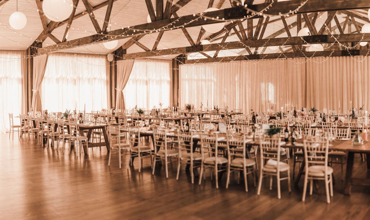 Wedding reception venue with rows of wooden tables and chairs arranged under draped fabric ceiling.