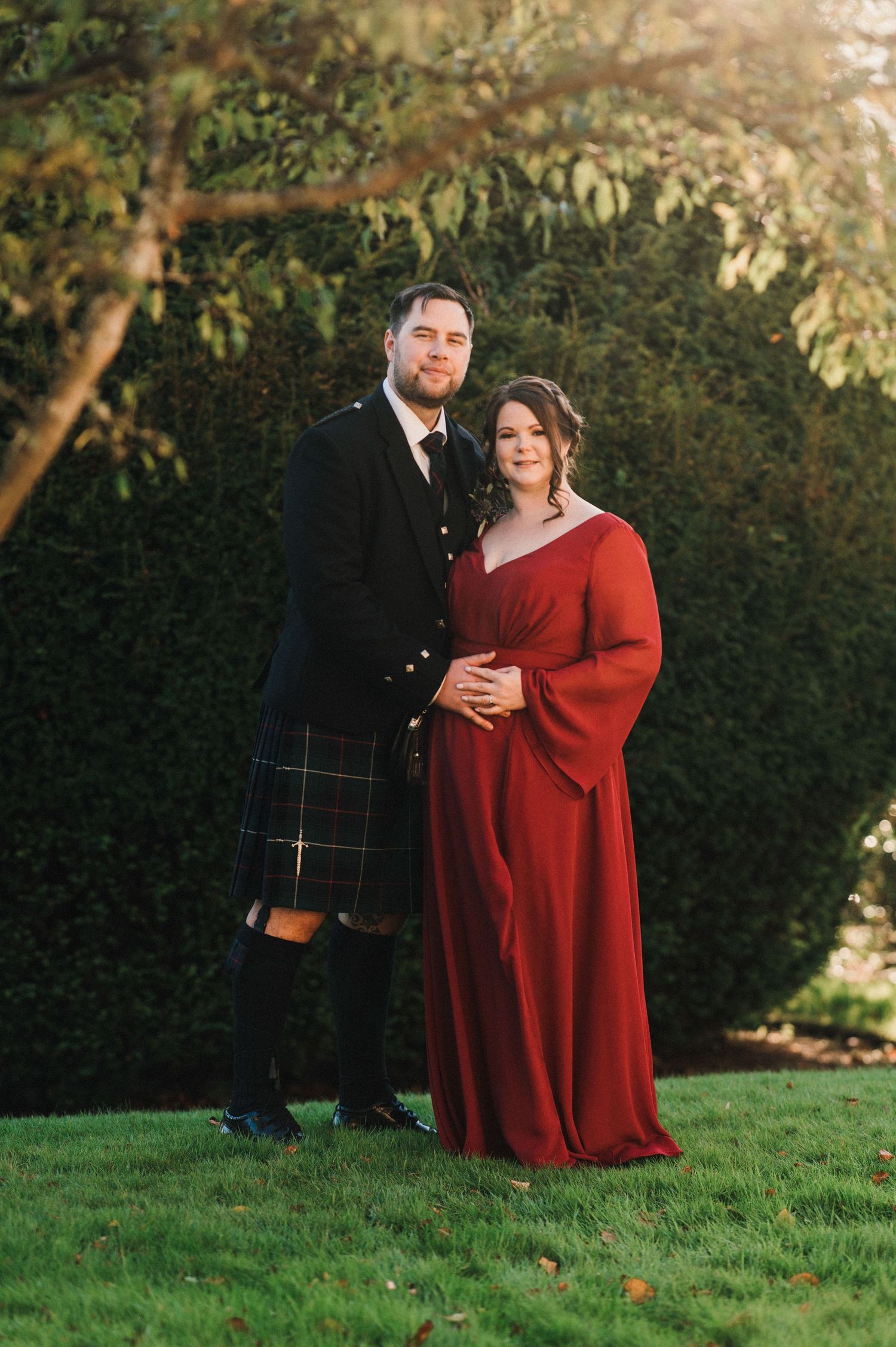 A formal couple poses outdoors, with one wearing a traditional Scottish kilt and the other in a flowing red dress.