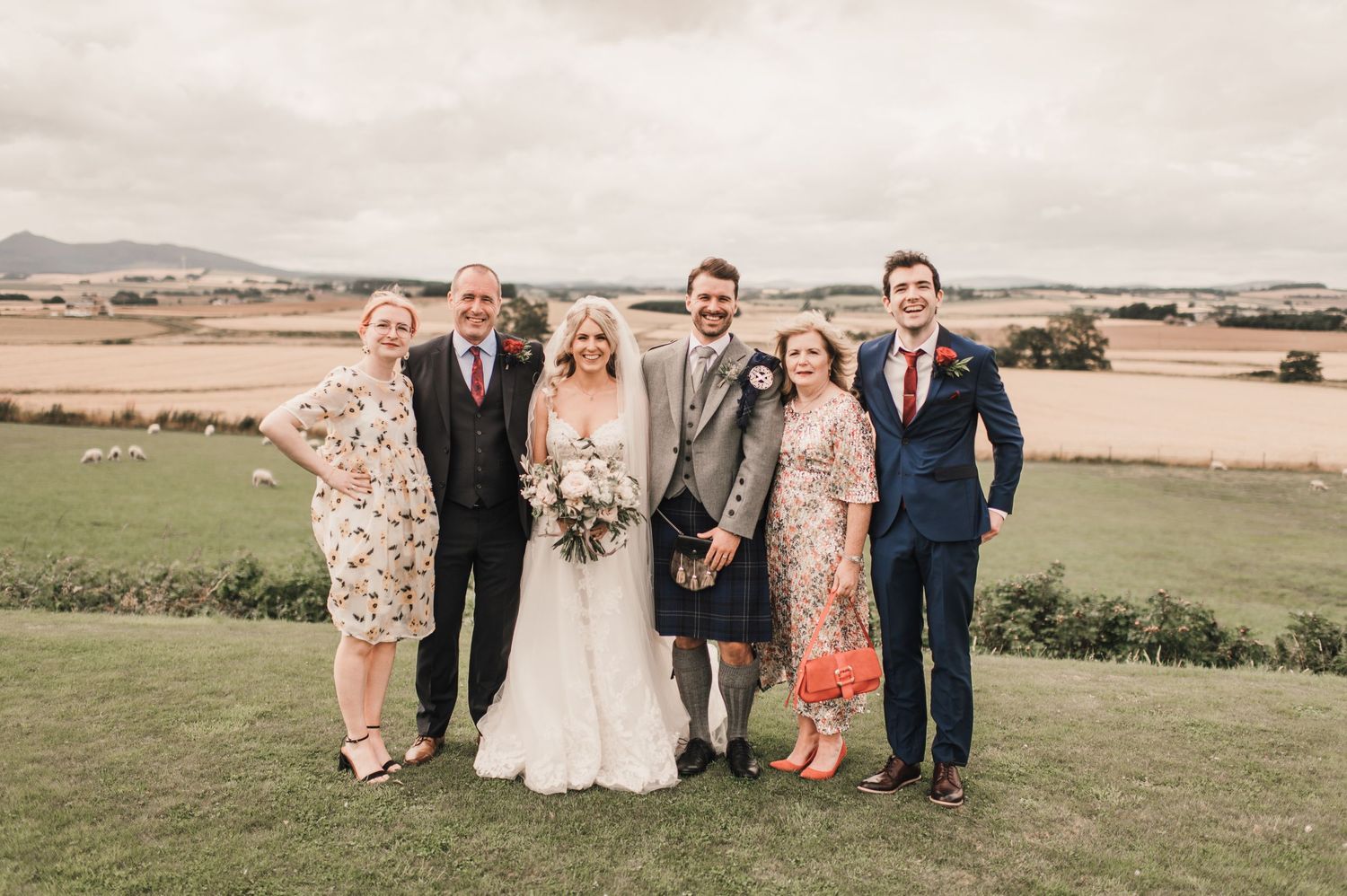 Small family group poses for formal wedding portrait on grassy hilltop with countryside views.