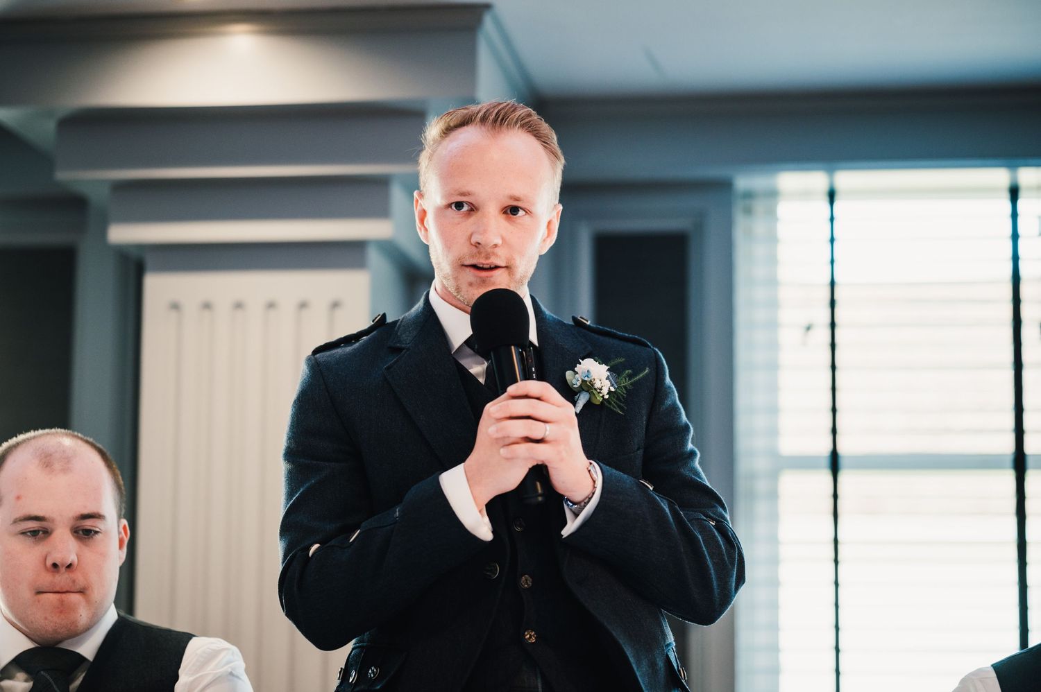 A wedding guest in formal attire delivers a speech using a microphone.