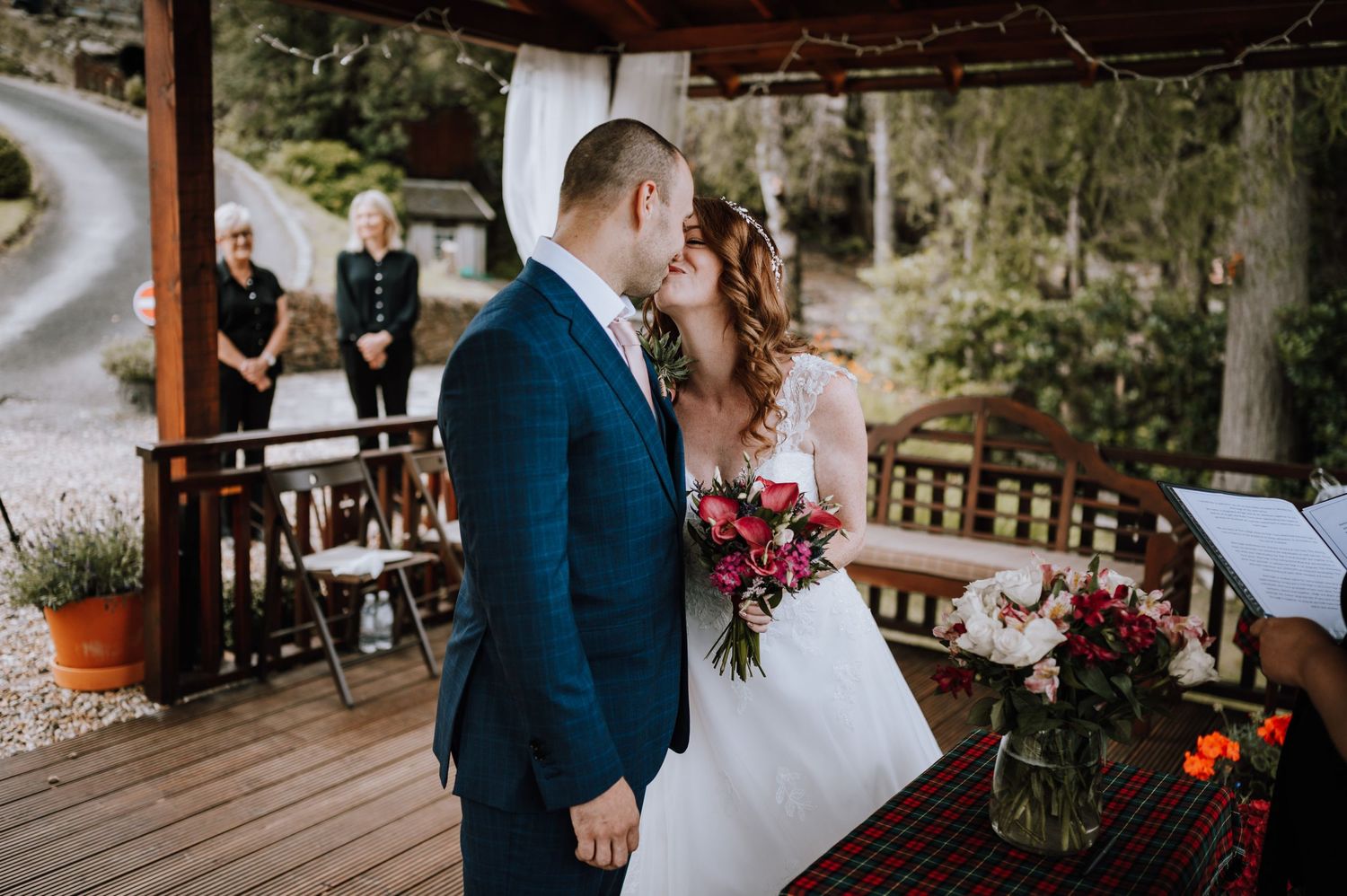 Couple exchanges vows under wooden pergola during lakeside wedding ceremony.