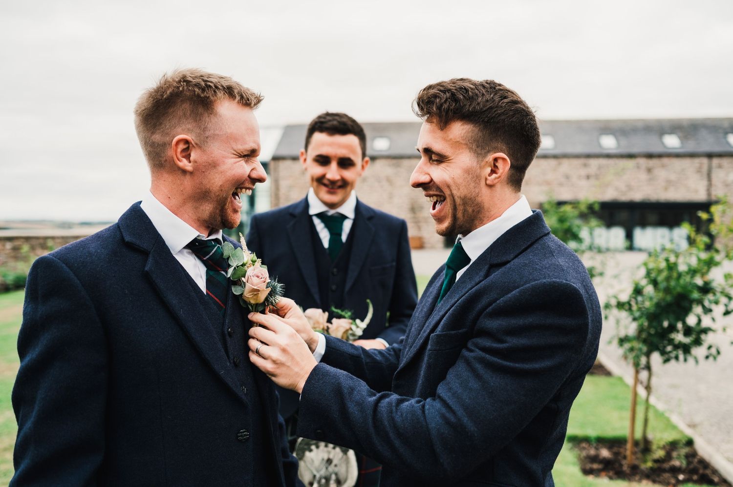 Wedding party members share a lighthearted moment while adjusting formal attire outdoors.
