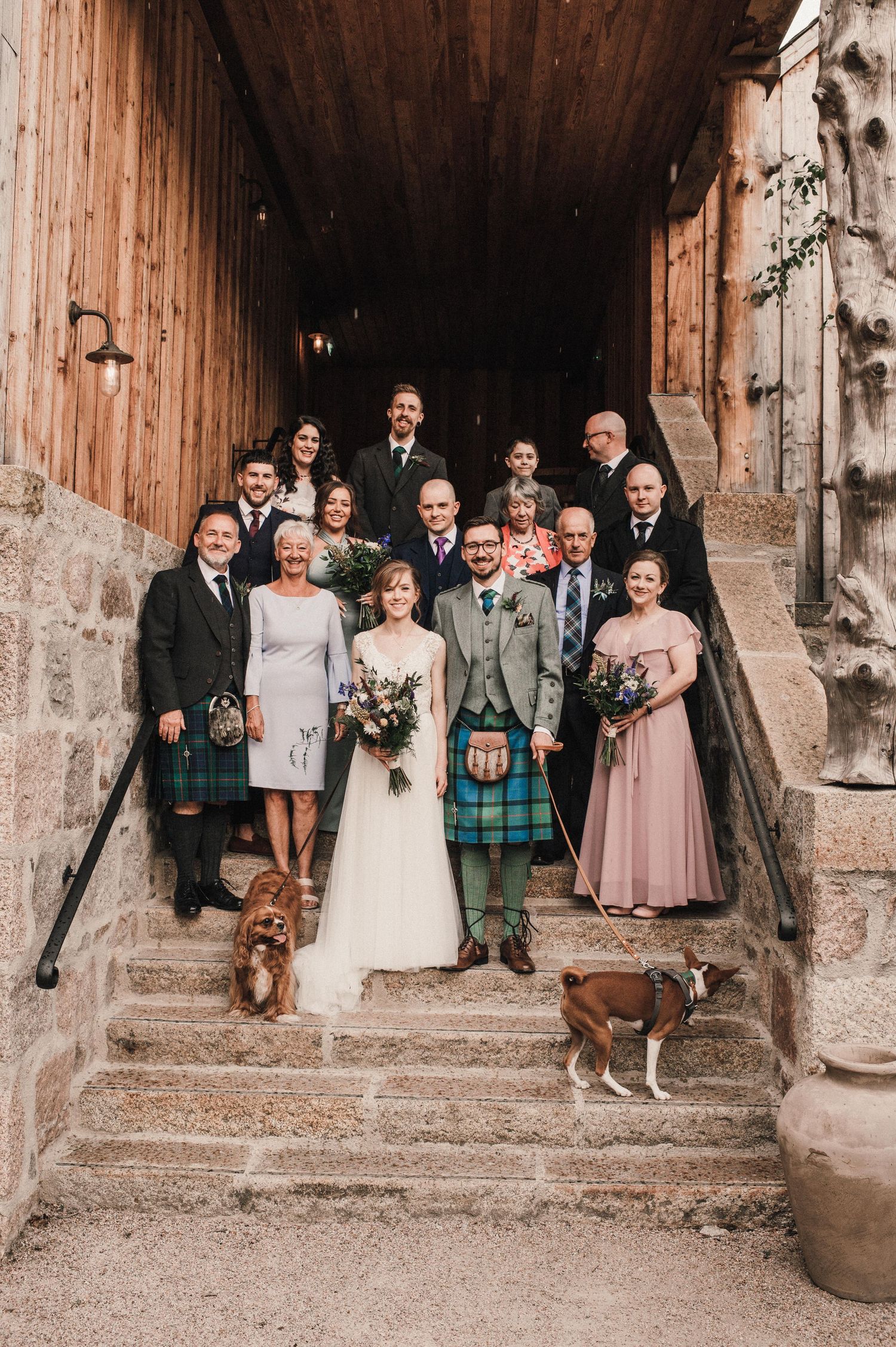 Large wedding party group photo on stone steps with dogs and traditional Scottish kilts.