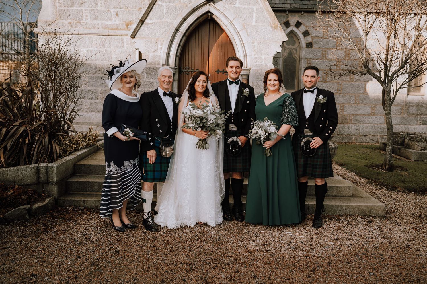 Wedding party in Scottish formal wear and kilts posed at church entrance.