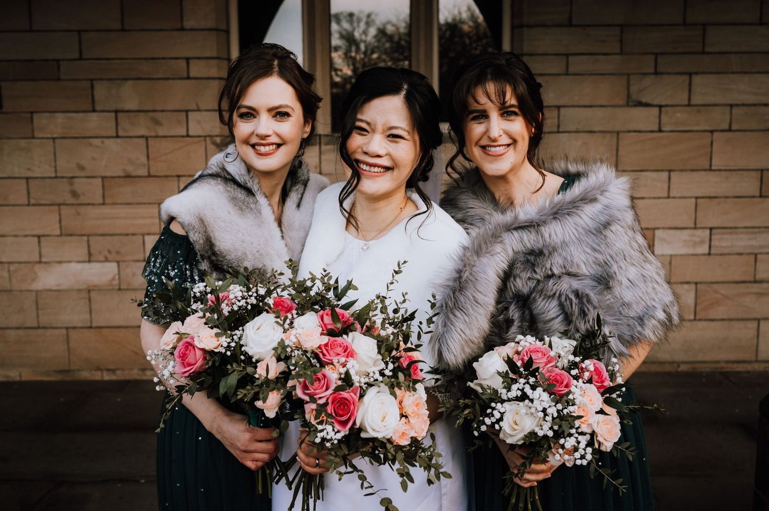 Three women in formal wear with fur shawls hold pink and white bouquets against a stone wall.