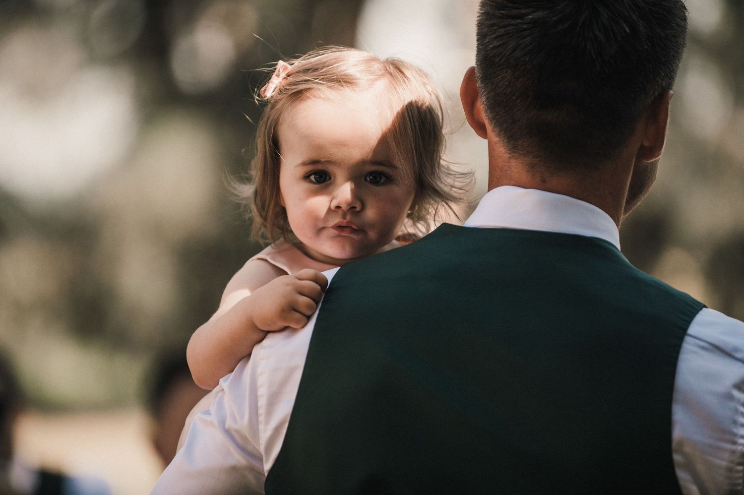 A small child peeks over someone's shoulder during what appears to be a wedding ceremony outdoors.