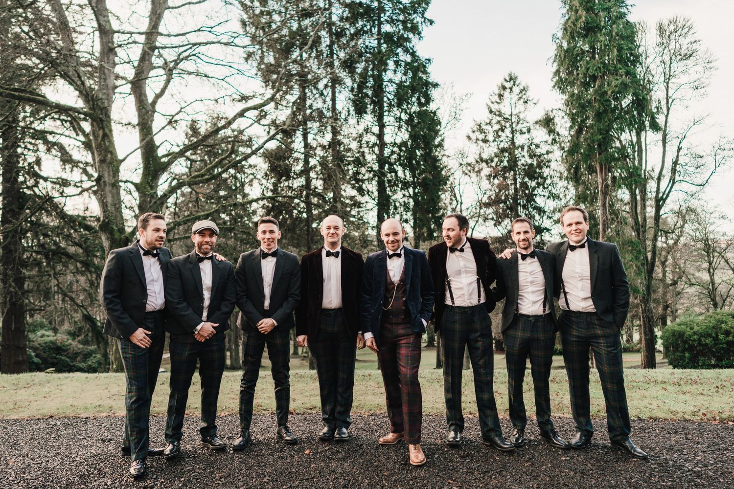 Wedding party of nine men in tuxedos pose together outdoors among tall evergreen trees.