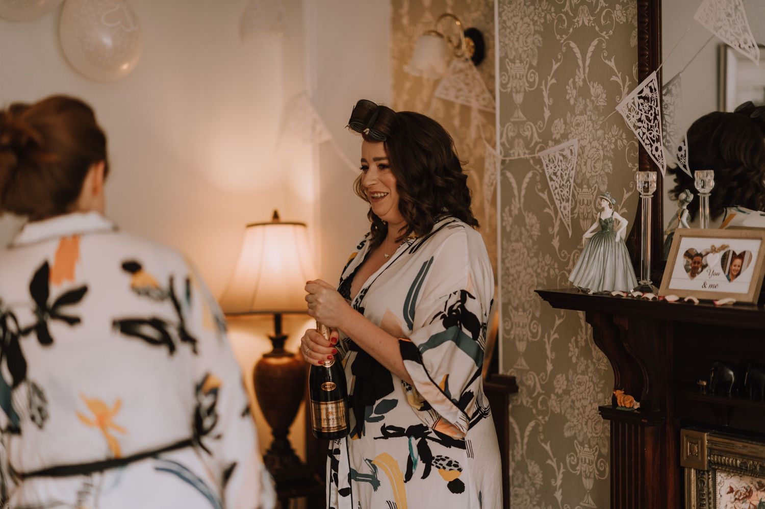 Two people chatting in living room with floral wallpaper and warm lamp lighting.