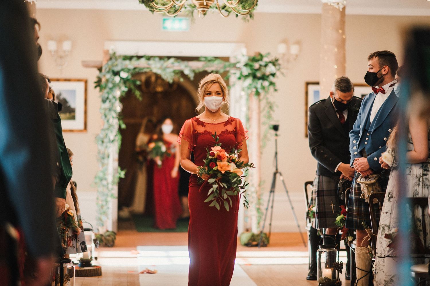 Bridesmaid in burgundy dress walks down decorated indoor aisle carrying autumn-themed bouquet.