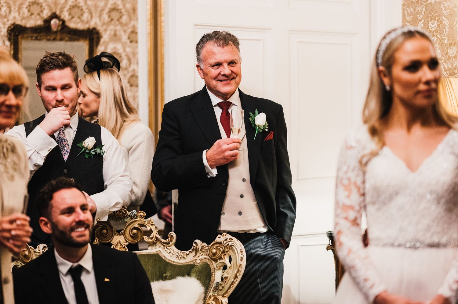 Wedding guests and speakers gather in an ornate room with decorative wallpaper during celebrations.
