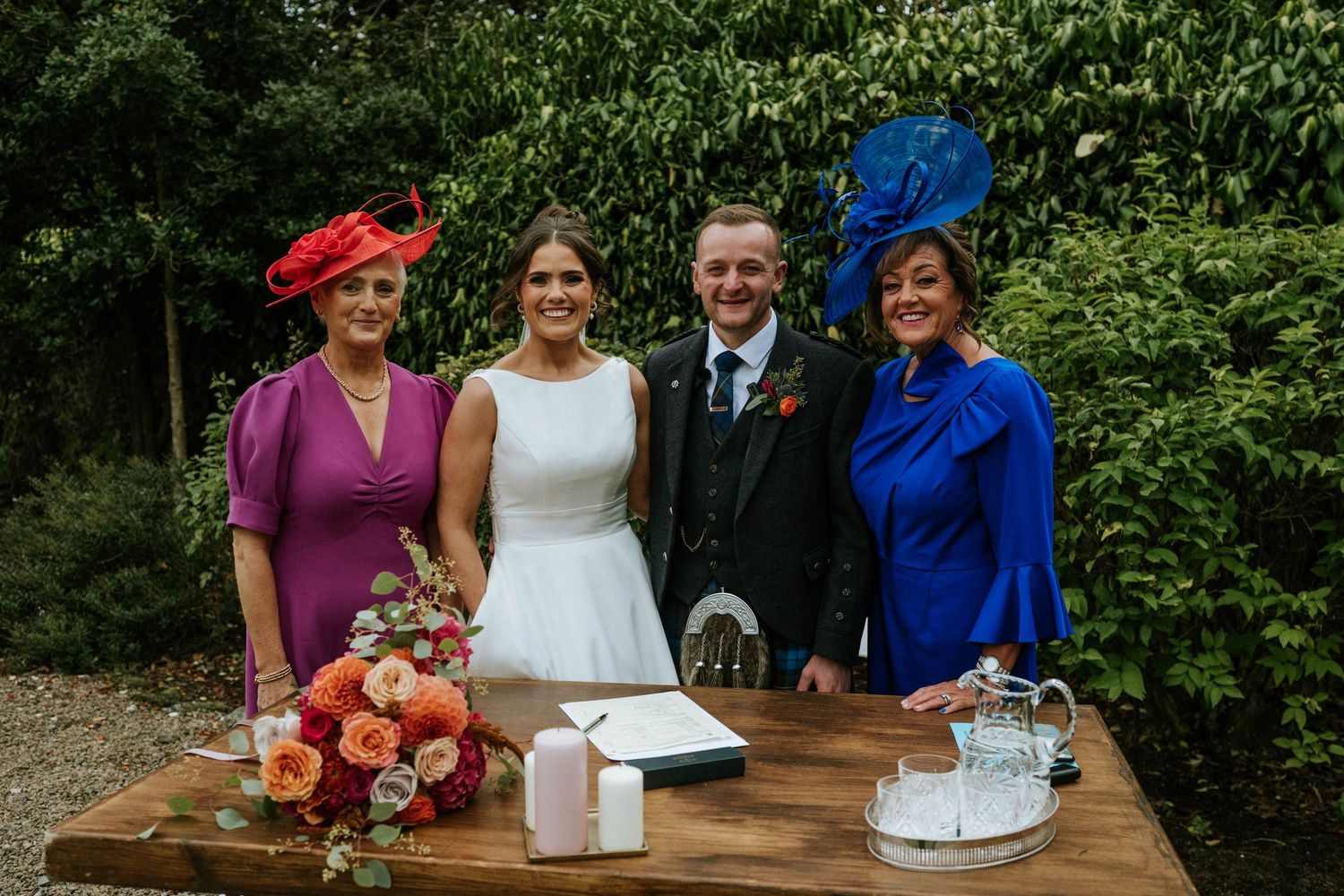 Wedding party gathered outdoors in front of lush green foliage wearing formal attire and traditional Scottish kilts.