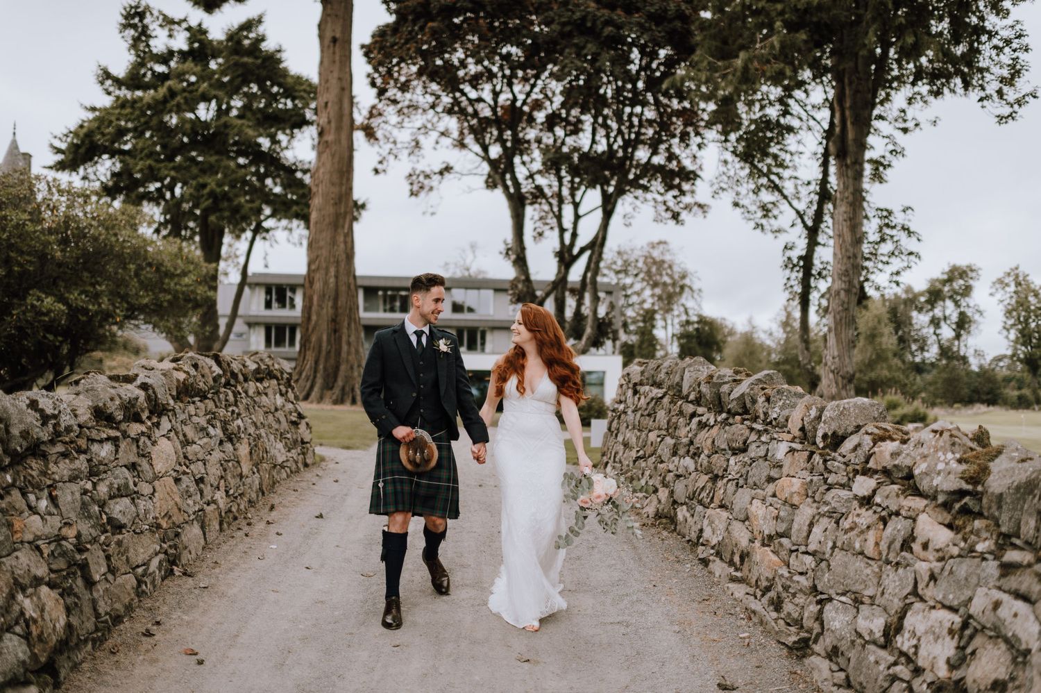 A couple walks hand in hand down a stone-walled path during their Scottish wedding.