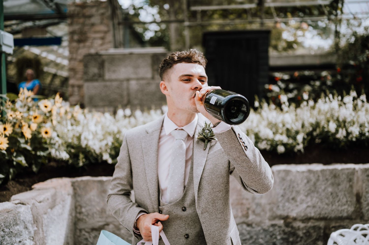 A guest in a light gray suit takes a drink during an outdoor wedding celebration.