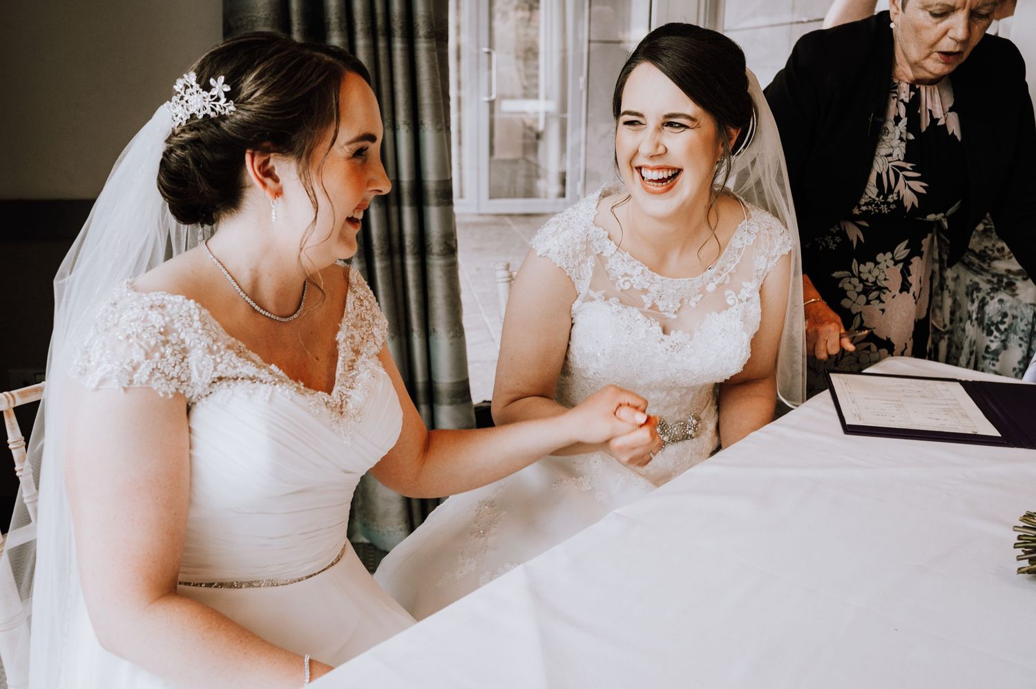 Two people in white wedding dresses share a joyful moment while signing documents at a white-clothed table.