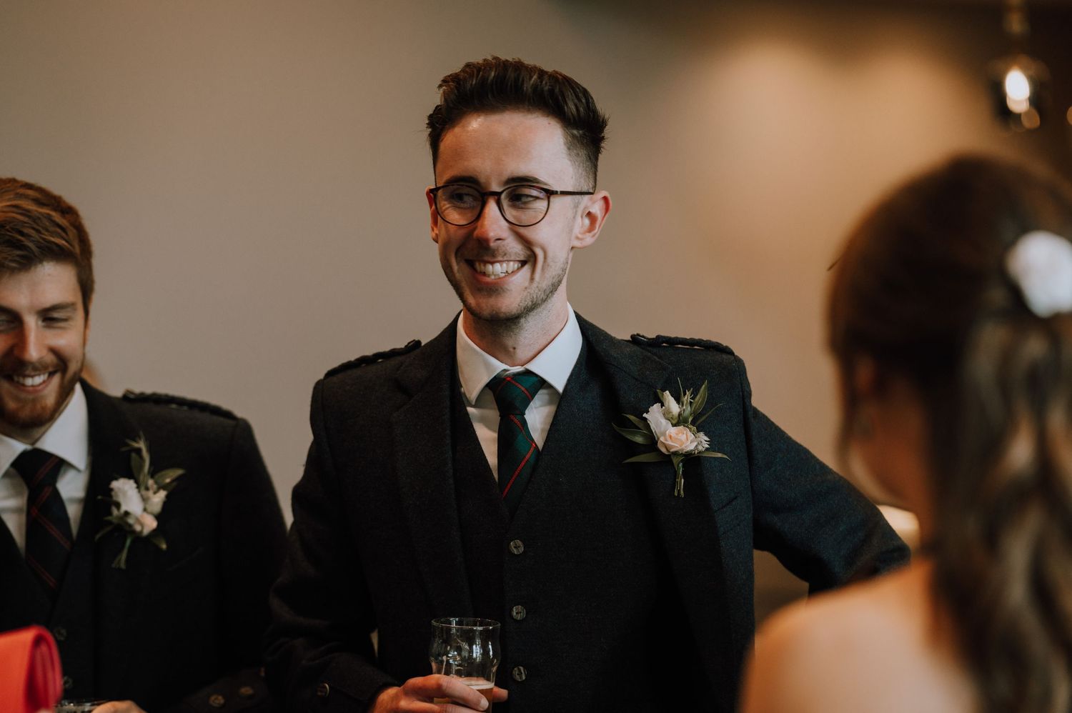 Wedding guests enjoying reception drinks in formal attire with boutonnieres at an indoor venue.