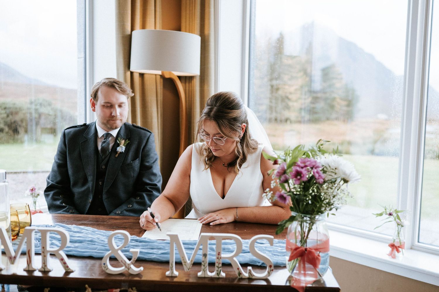 Couple sits at a bright window-lit table during a wedding reception with white and purple floral centerpieces.