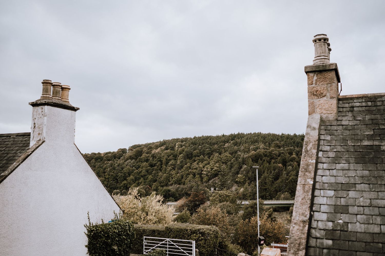 White cottage houses with chimneys against cloudy sky and forested hillside.