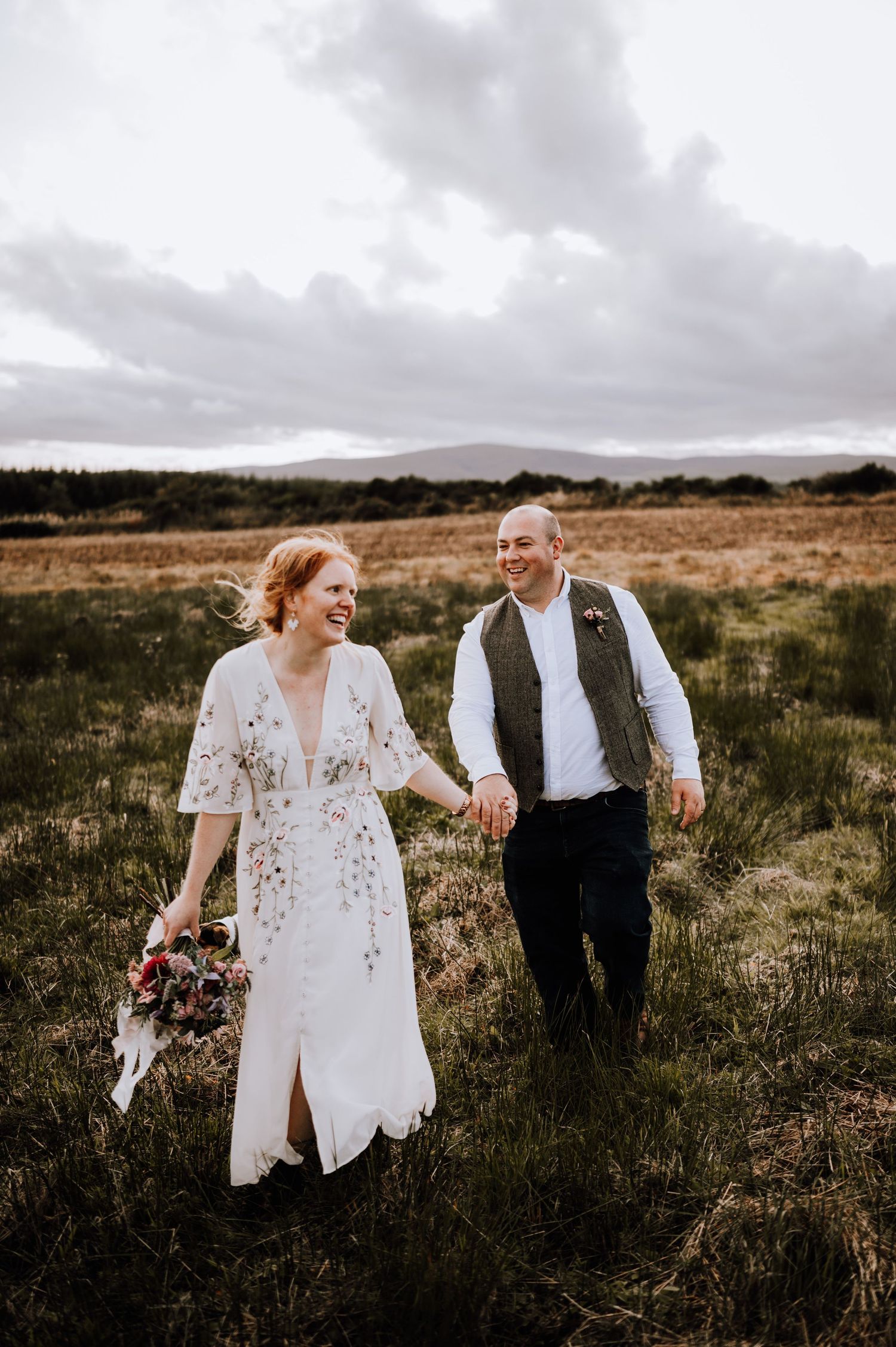 A couple walks hand in hand through a rustic field with mountains and cloudy skies in the background.