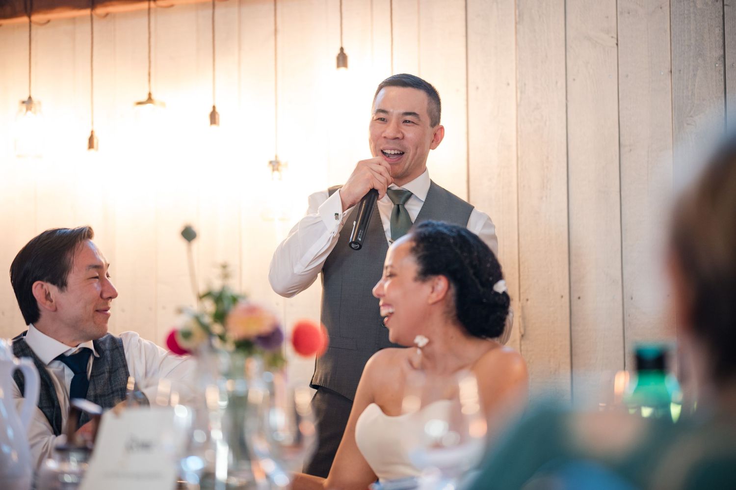 Indoor wedding reception with hanging lights and guests celebrating at decorated tables.