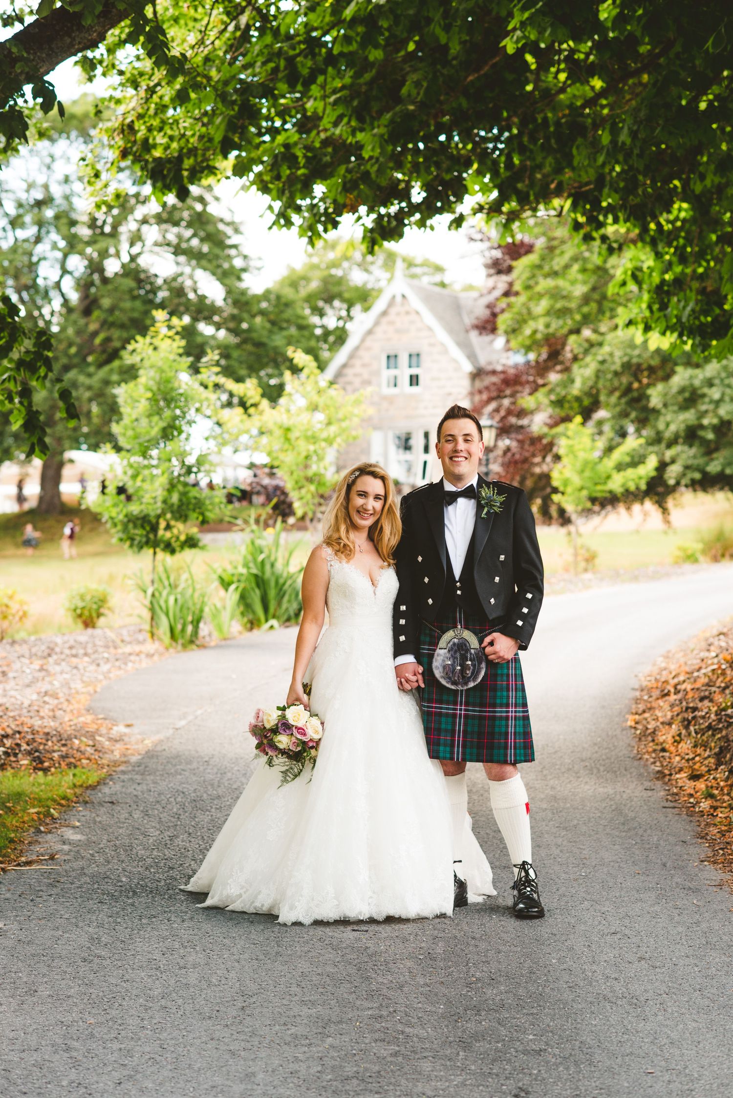 Wedding couple walking along a tree-lined path with traditional Scottish kilt attire.