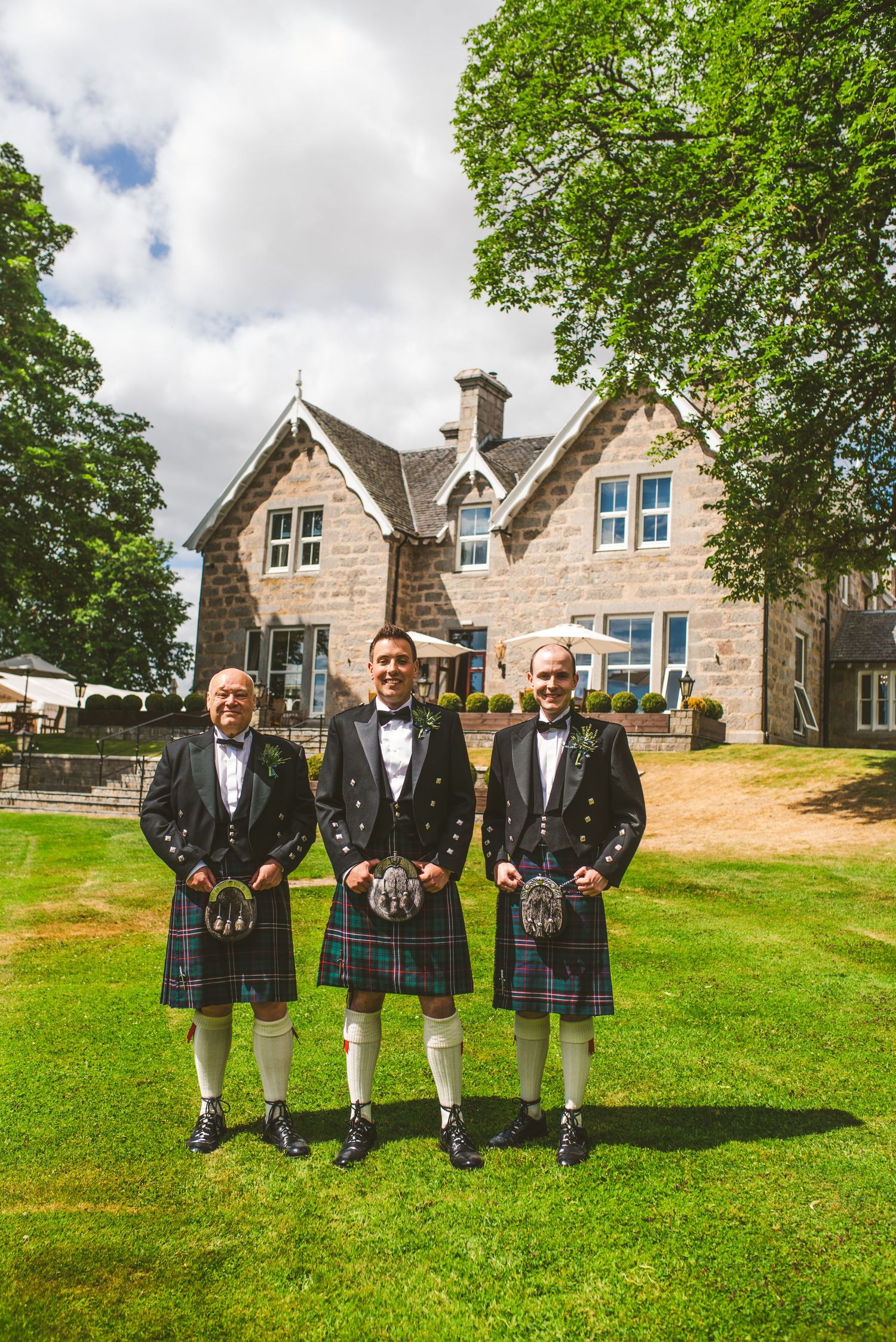 Groomsmen wearing traditional Scottish kilts and formal jackets pose on a green lawn with stone buildings in background.