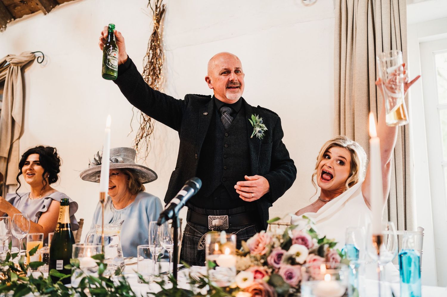 Wedding guests celebrate and laugh during reception speeches at wooden-paneled venue.