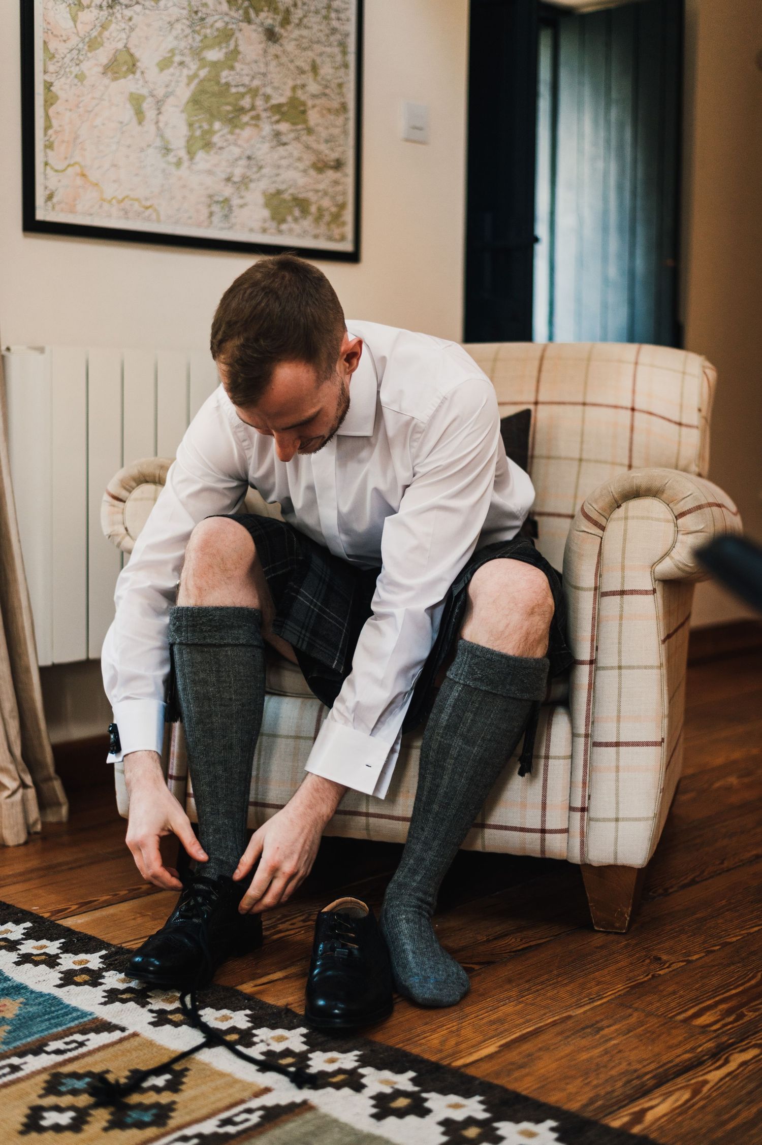 Person in formal attire tying black dress shoes on hardwood floor with white furniture in background.