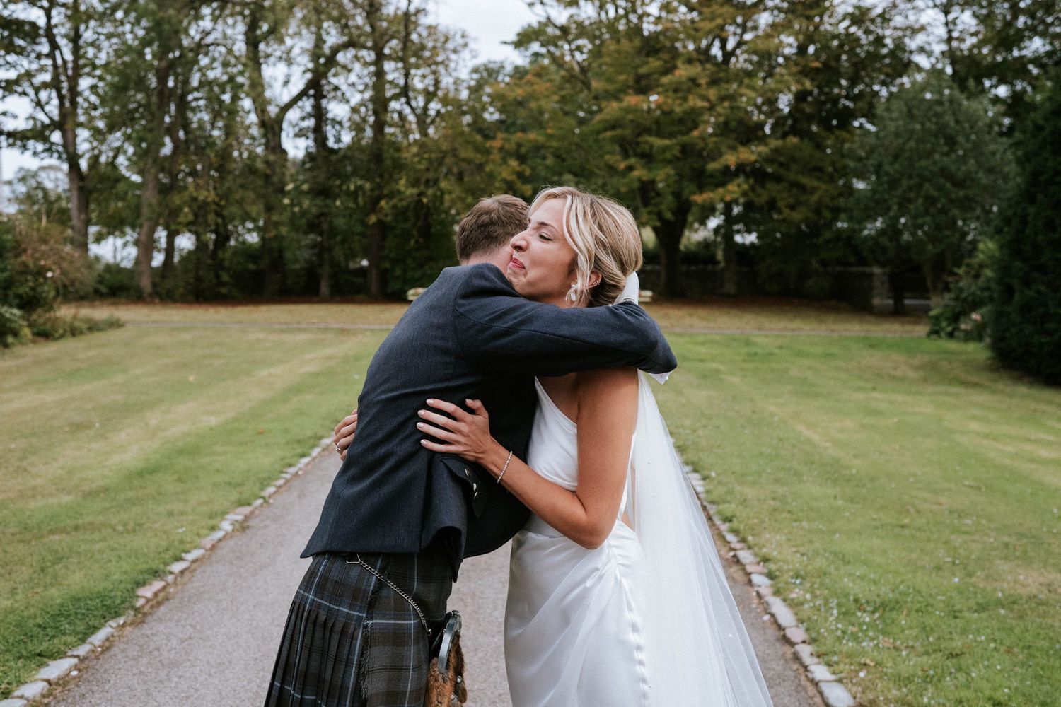 Couple embracing on a scenic path surrounded by green lawn and trees.