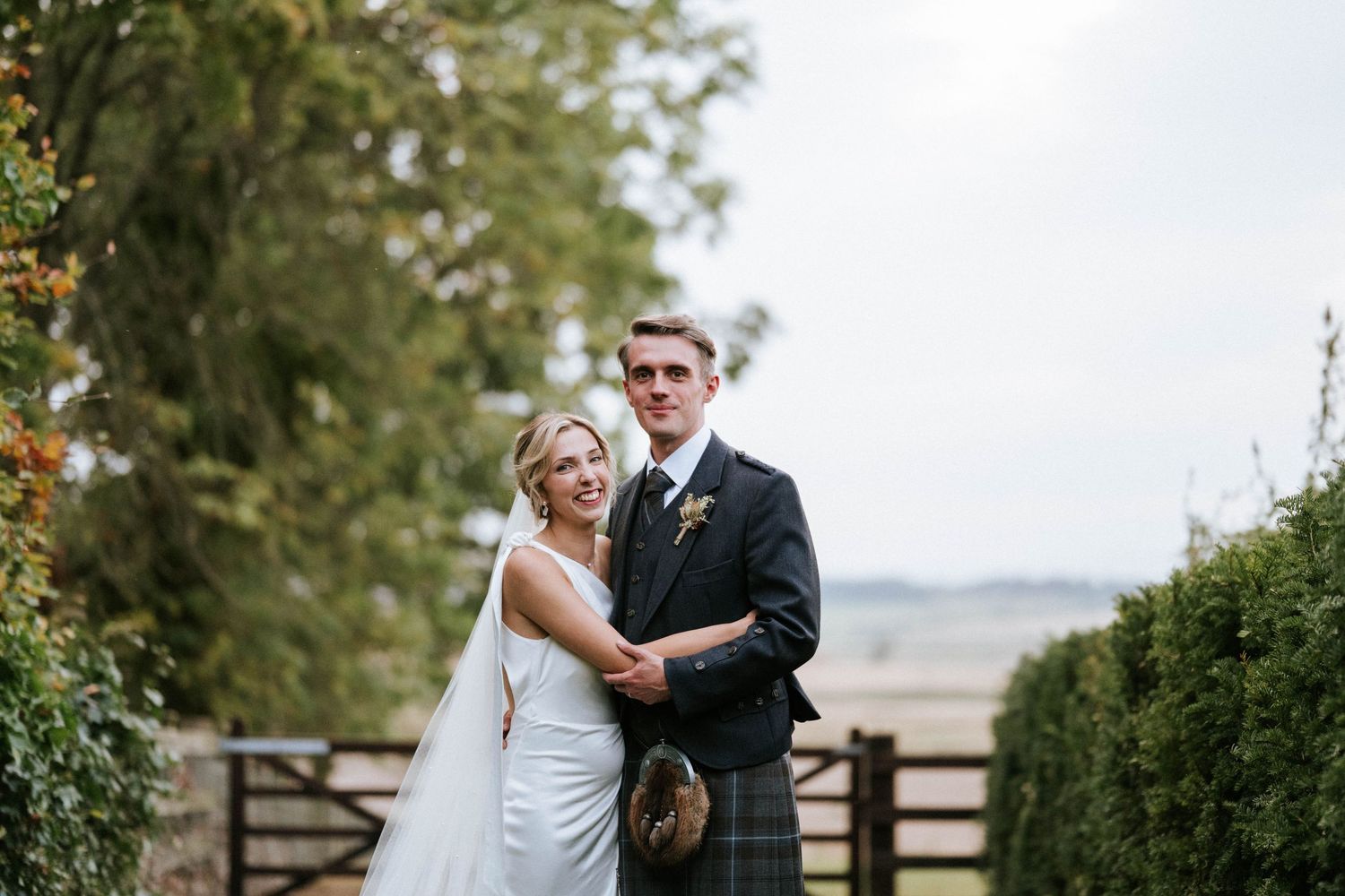 A couple poses together in wedding attire against a countryside backdrop.