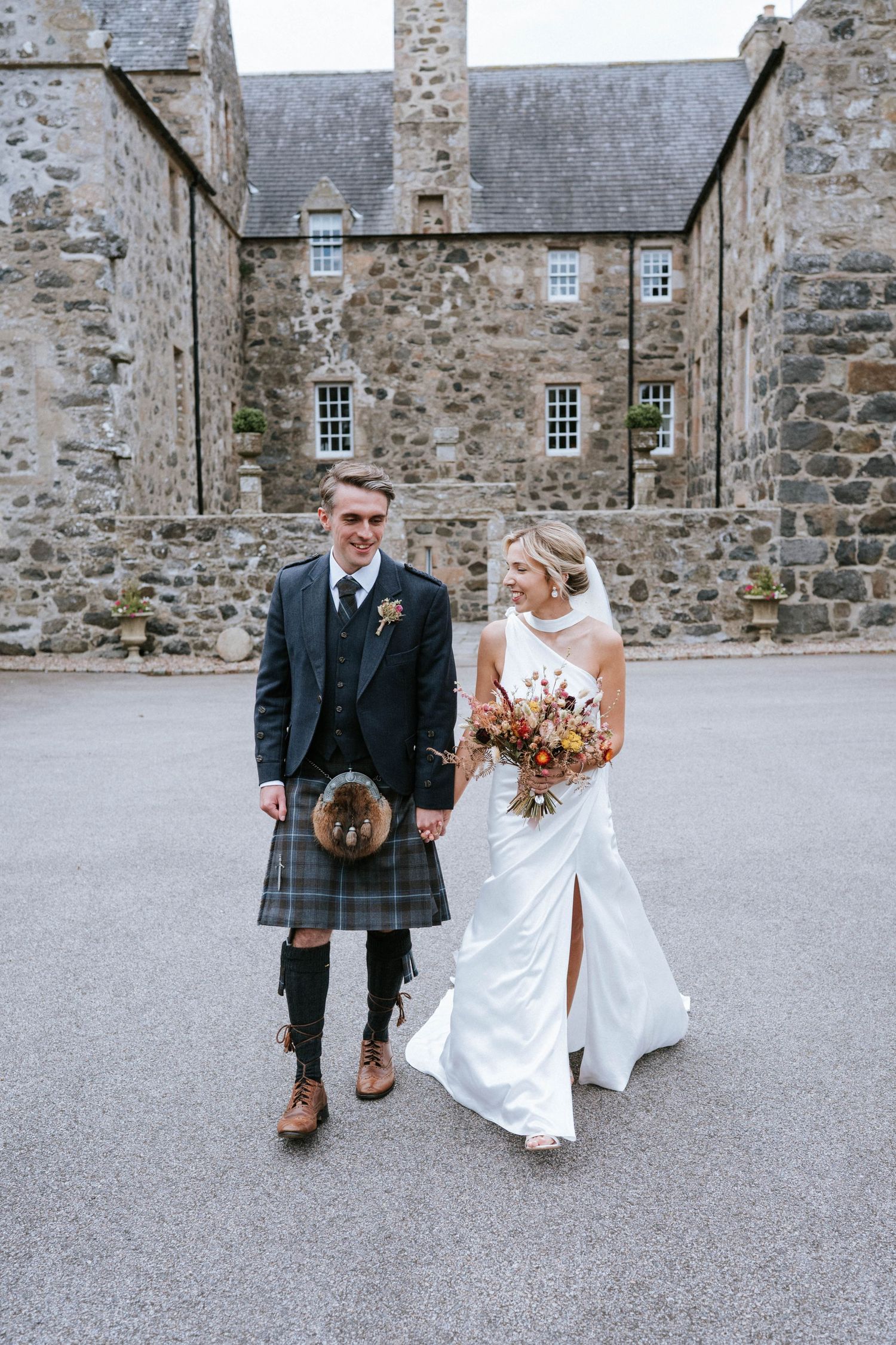 Newlyweds pose playfully outside a historic stone castle building in Scotland.