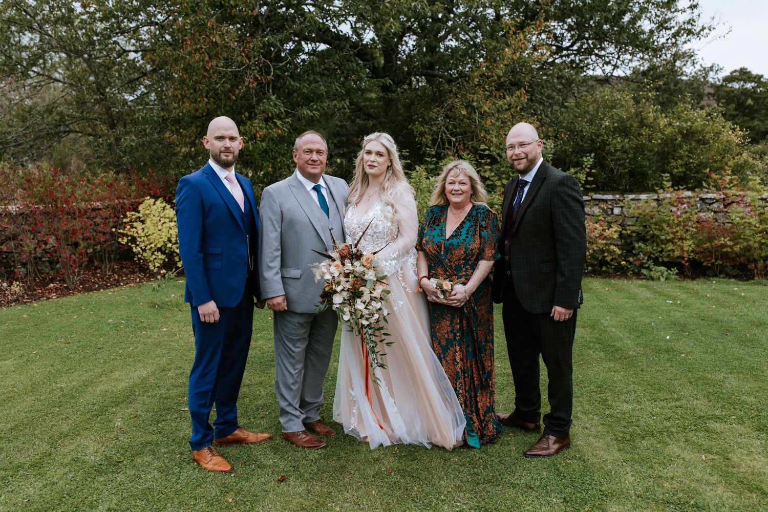 A wedding party poses together on a manicured lawn with trees in the background.