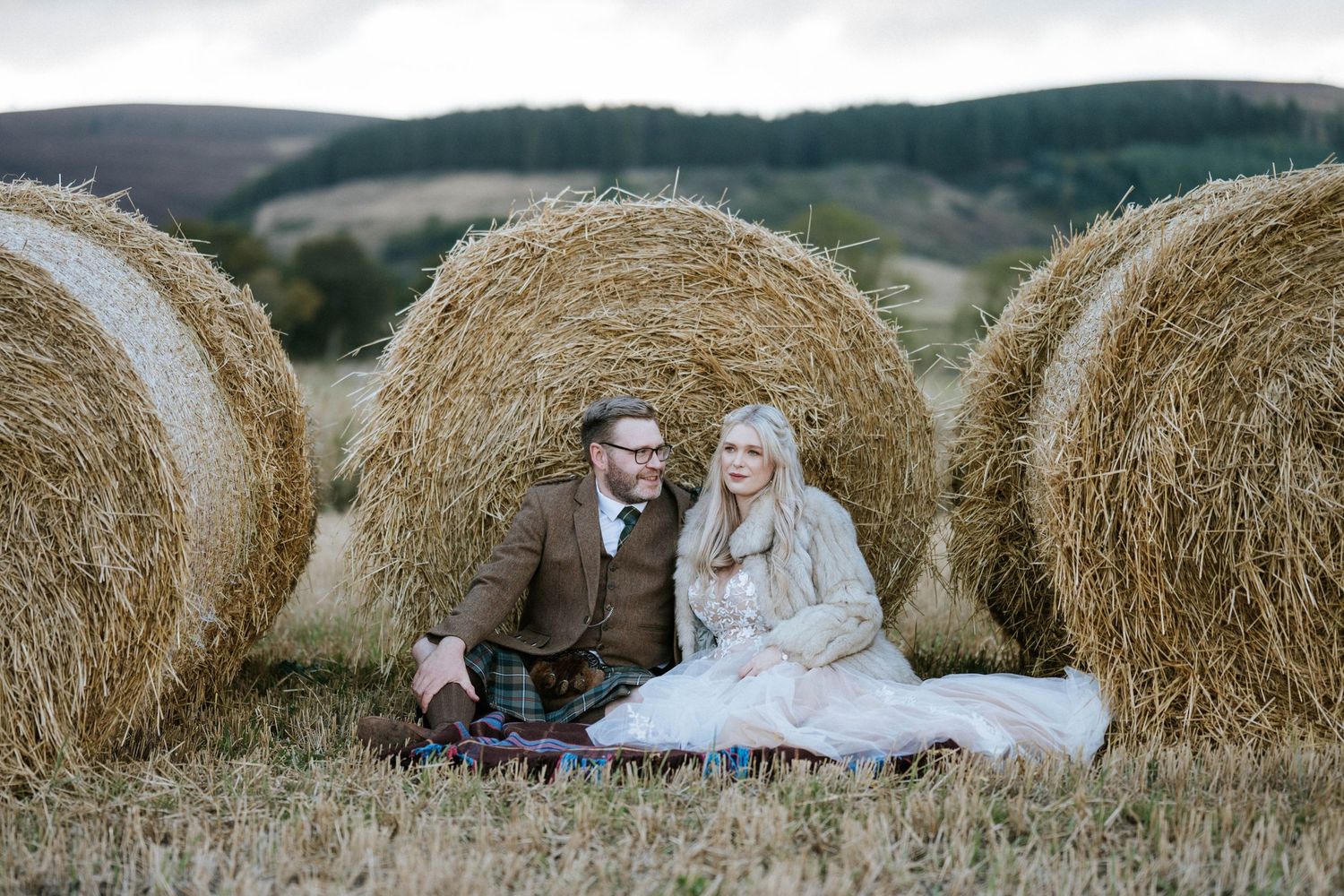 Newlyweds share an intimate moment among round hay bales in a Scottish field.