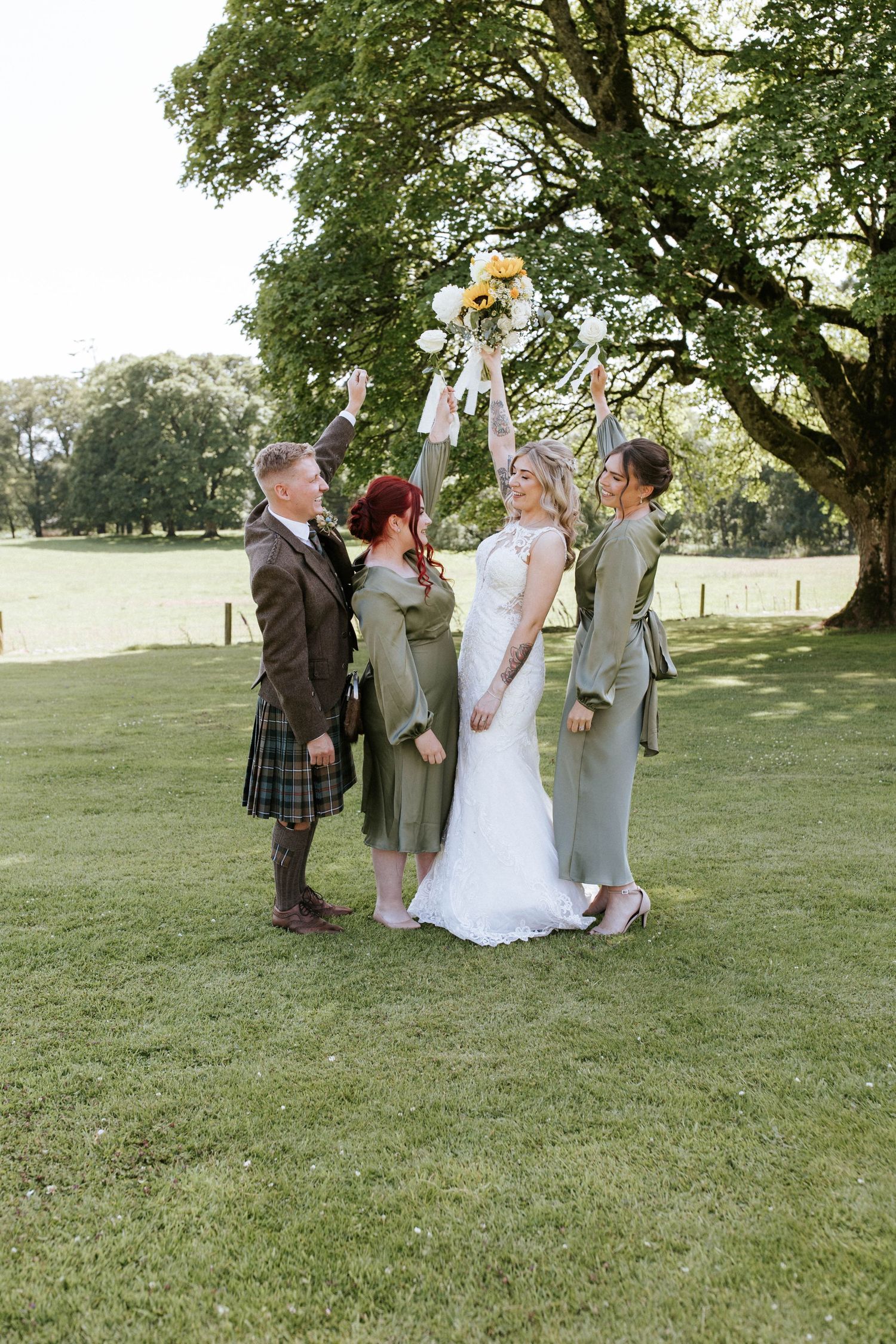 Wedding party celebrating outdoors under a large tree on a sunny day in Scottish-inspired attire.