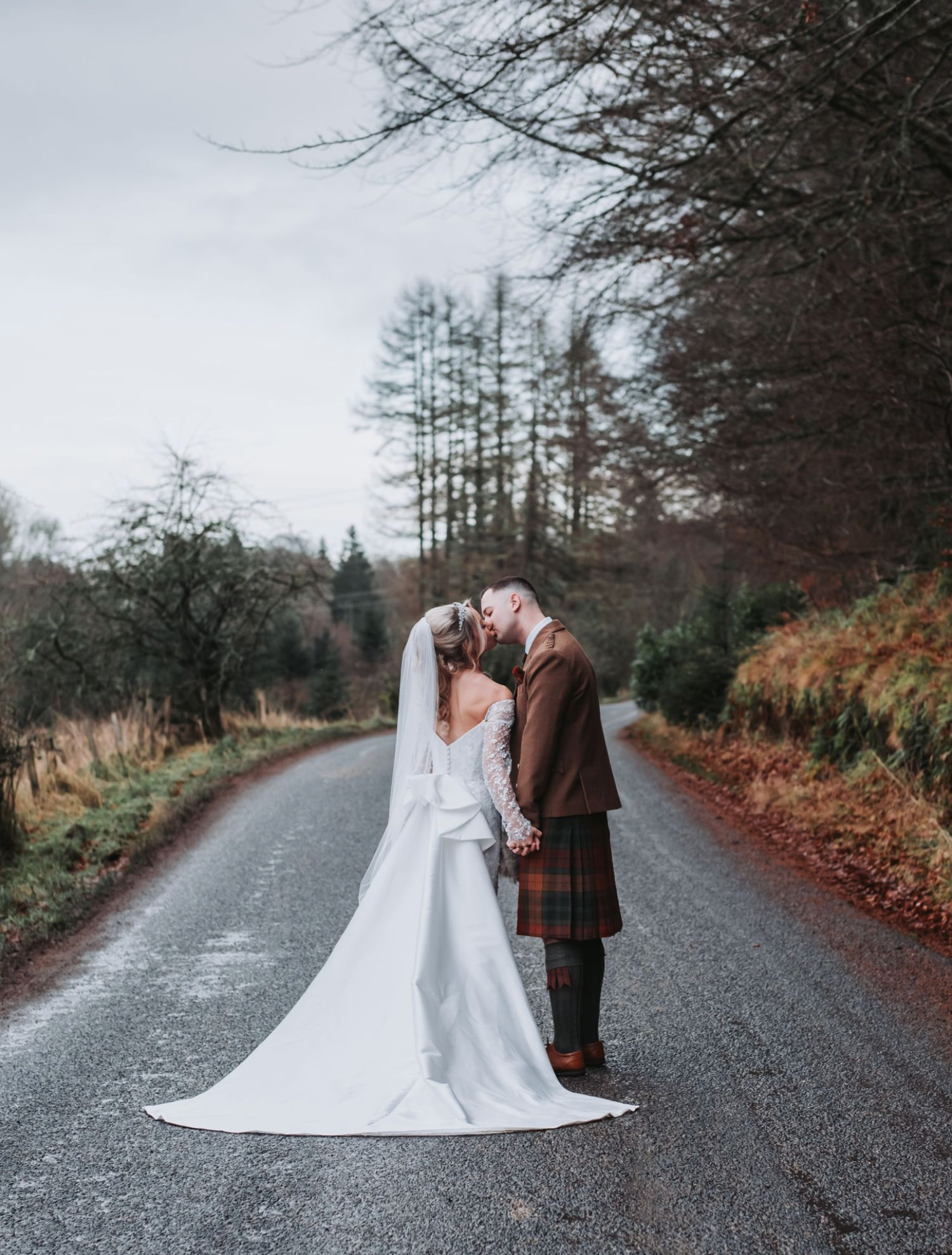 A couple in wedding attire poses together on a rural road surrounded by autumn foliage and evergreen trees.