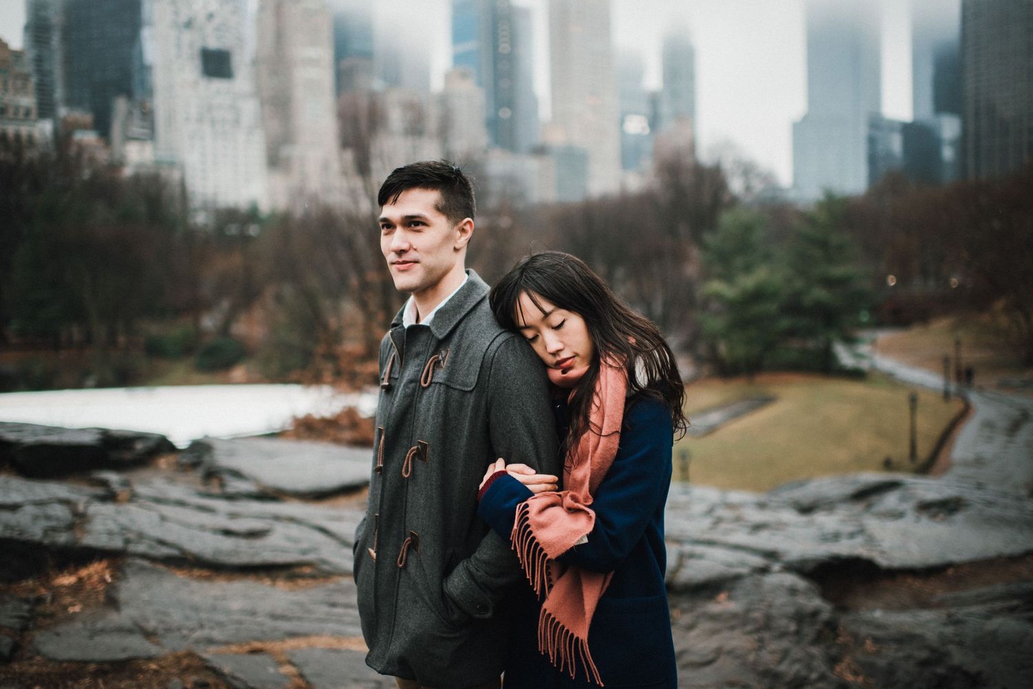 A couple stands together in Central Park with the Manhattan skyline visible behind them on an overcast day.